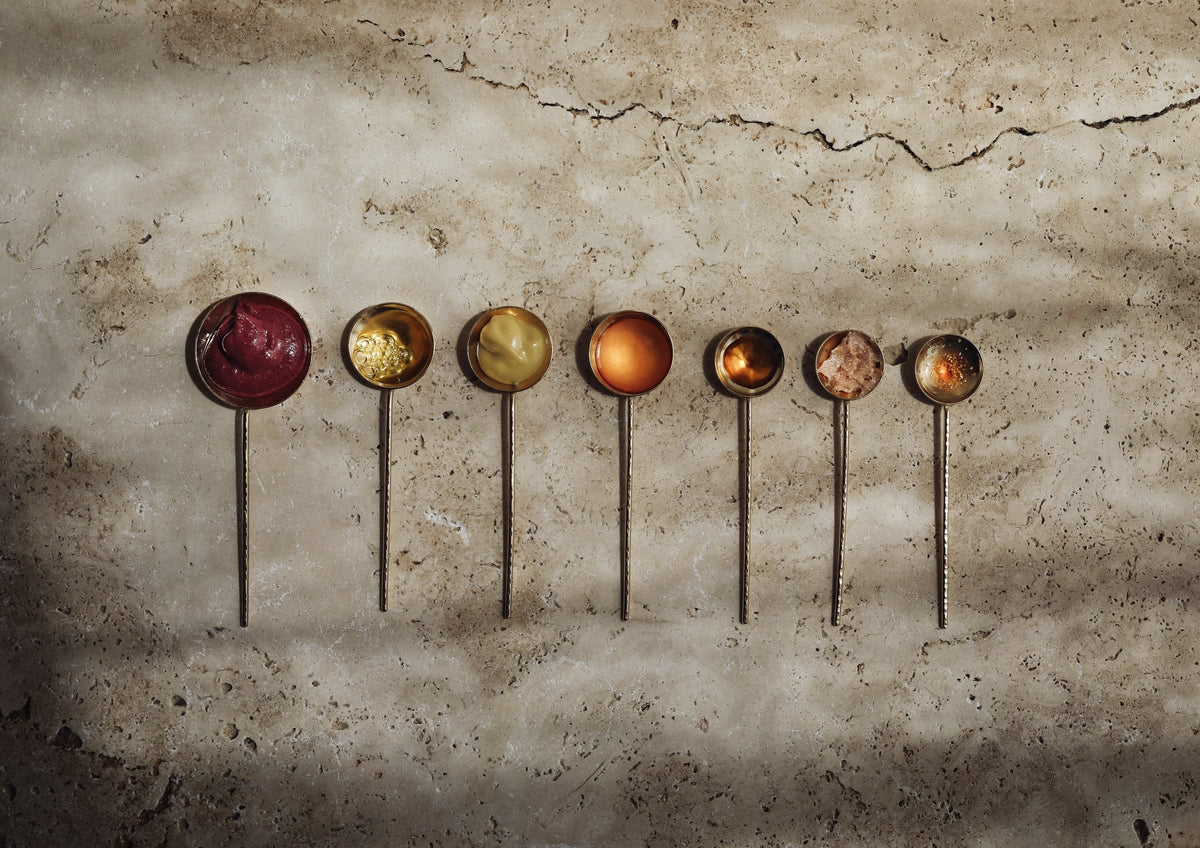 seven brass ladles with balms and oils on pale stone